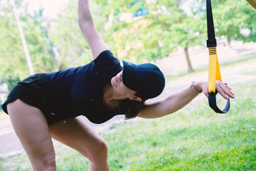 A focused woman in a black cap and workout gear performs an outdoor exercise with a TRX suspension trainer in a vibrant green park, highlighting fitness and an active lifestyle.