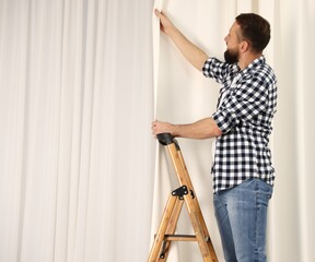 Man with ladder hanging up window curtains at home, space for text © New Africa
