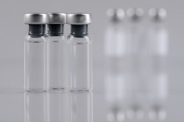 Vaccine in glass vials on mirror surface against blurred grey background, closeup. Space for text