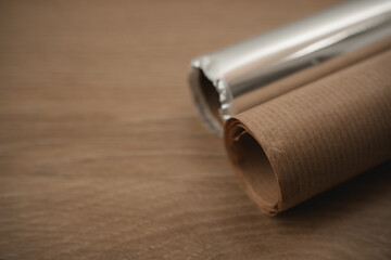 Two rolls of gift wrapping paper are placed on a wooden table, ready for wrapping presents for Christmas. One roll is brown kraft paper, and the other is shiny silver foil.