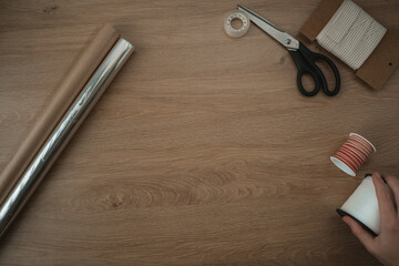 A person prepares to wrap presents on a wooden table. Scissors, tape, and gift wrapping materials are arranged neatly. The festive spirit is evident with holiday-themed decorations nearby. Copy space 