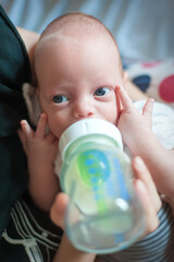 A baby holds a bottle with both hands, focusing on the flow of milk. The caregiver is nearby, creating a warm and nurturing environment in their home living space.
