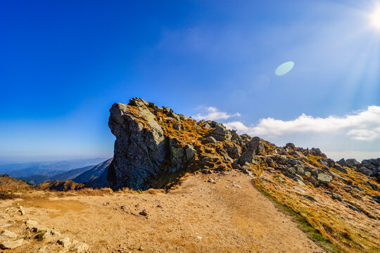 A wide panoramic view of a rocky hiking trail winding through the slopes of the Low Tatras National Park during a sunny autumn day. Beautiful yellow grass and blue sky