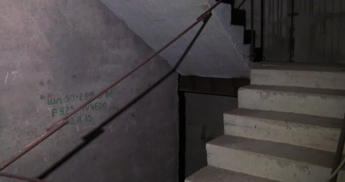 a slow, descent down a rugged concrete stairwell within an unidentifiable, dark structure. The camera steadily pans, revealing concrete steps, a metal railing, and cryptic markings on the grimy walls