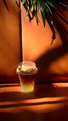 Iced beverage in a clear plastic cup placed against a warm-toned background with natural sunlight and tropical leaf shadows. The contrast of light and shadow creates a modern, relaxed mood, suitable f
