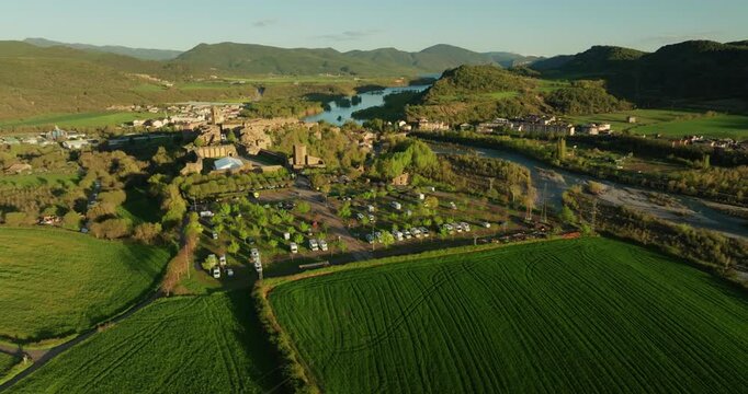 High-altitude drone orbit over irrigated farmland and caravan park in the Cinca valley with views toward A&iacute;nsa and the Pyrenean foothills of Sobrarbe, with large rectangular cereal fodder crop fields