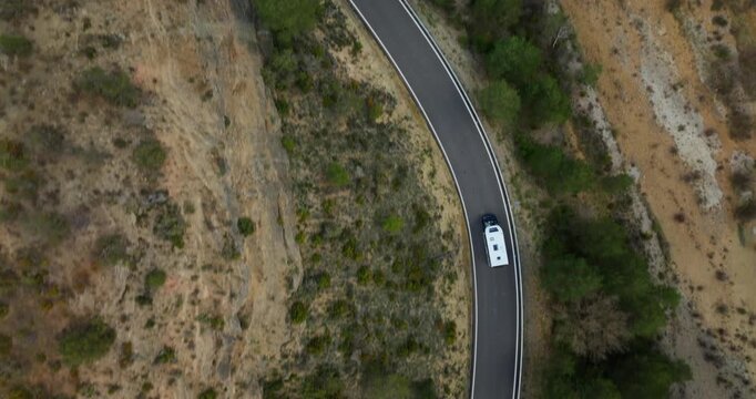 Overhead drone follows a white camper van traveling down a narrow dark asphalt road cutting through sparse Mediterranean shrubland with open ground typical of Spanish rural countryside.