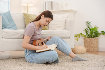 Young Asian woman sitting on carpet in living room writing lyrics in notebook while playing ukulele composing music and relaxing at home