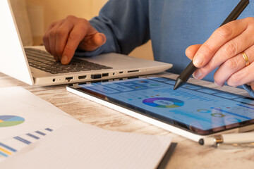 Person analyzing financial data on tablet with stylus while typing on laptop, calculating taxes, managing online payments and budgeting with charts and documents on a home office desk