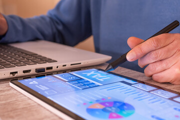 Person's hand using a tablet tax planning dashboard with charts, numbers and tax visible, laptop in background illustrating digital finance, accounting and online tax management