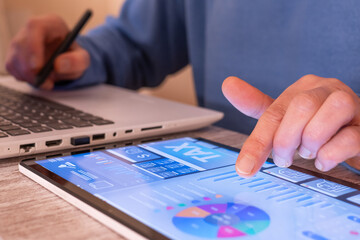 Person interacting with a digital tablet displaying a tax planning dashboard and financial data while also working on a laptop, calculating and managing online finance using modern technology