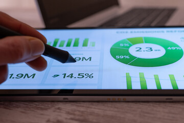 Hand holding a stylus pen, interacting with an esg dashboard displaying green charts, graphs, percentages, and metrics for corporate responsibility and sustainable business development
