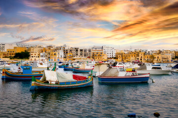 View of Birzebbuga's small harbor with numerous moored boats at sunset ; rows of houses appear in the background.