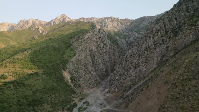 Aerial view of a narrow canyon carved between steep cliffs in Uzbekistan during early morning. Soft sun glows on green slopes and stone wall, the road and dry stream guide the eye through depth