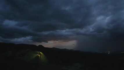 Dramatic zoom out timelapse of couple camping in tent top mountain thunderstorm brews, glowing shelter against dark clouds and lightning in distant horizon.Concept hikers under lightning sky isolated