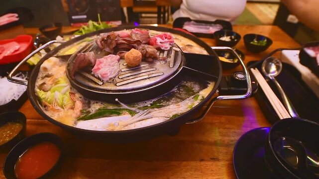 Top view of traditional Thai BBQ hotpot (Moo Krata) with mixed seafood, octopus, shrimp, fish balls, noodles, mushrooms, cabbage, corn, garlic, and dipping sauces on wooden table in Bangkok, Thailand.