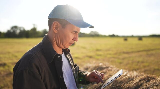 Man in field with tablet. Farmer working on field with haystacks and tablet. Farmer working in a field with haystacks of grass. A man standing in a field holding lifestyle a tablet.