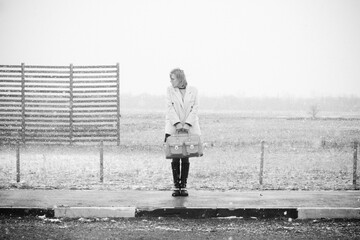 Young woman in a gray coat holding a large stylish bag standing on a road during a snowfall. Winter fashion and lonely travel concept.