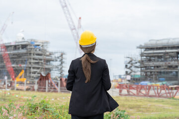 An Asian female engineer wearing a safety helmet stands with her back turned, observing the company's industrial plant to support business planning, machine control, and sustainable development.