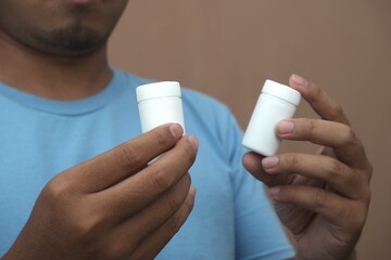 Close-up of Southeast Asian man holding two white pill bottles, representing medication choice, healthcare decision, and pharmaceutical concept for medical and advertising use.