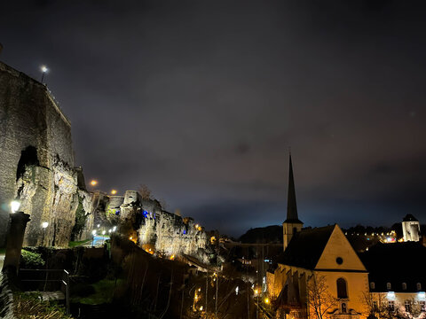 Night view over the Pfaffenthal valley in Luxembourg City, featuring illuminated rocky fortifications and a historic church with a tall spire. Warm city lights contrast with dark cliffs and a clouds