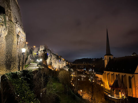 Night view of the Pfaffenthal valley in Luxembourg City, with illuminated rocky fortifications and a quiet path running along the cliffs. A historic church with a tall spire rises on the right side