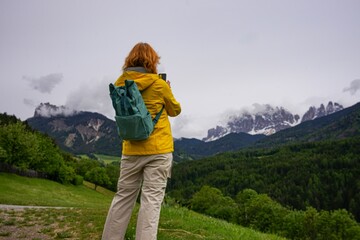 Woman with backpack taking a photo of mountain landscape using smartphone. Female hiker standing outdoors in green alpine valley with forest and rocky peaks in the background.
