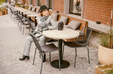 Business woman in grey suit sits on outdoor cafe terrace on autumn day, adjusting hat with warm confident smile, in scarf and gloves, relaxed city moment. Business lunch, urban lifestyle concept.