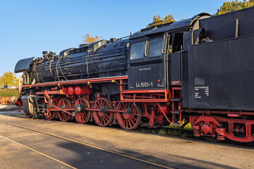 Detailed view of the large red driving wheels and mechanical rods of a black vintage steam locomotive resting on the tracks in sunlight.