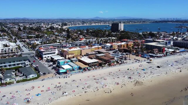 Pacific Beach in San Diego, California. Aerial drone showing a crowded sandy shoreline, beachfront buildings, and Mission Bay on a clear sunny day. Downtown SD buildings on distant skyline.