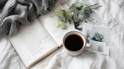 A cozy scene with an open book, coffee cup, leafy sprigs, and photos resting on white linens, alongside a folded knitted blanket