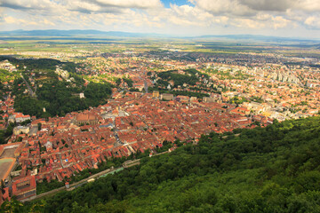Fototapeta premium View from hill showing old and new Brasov with red rooftops, cityscape merging with green hills on a sunny day.