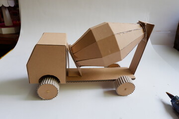 A brown cardboard model of a cement mixer stands on a white surface. The construction model features a detailed cement drum, wheels, and base. Neutral background 
