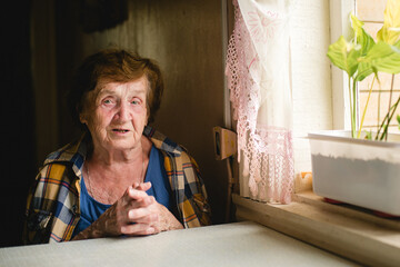 Elderly woman sitting by a window in a modest home, softly lit by natural light, hands clasped, with a calm, thoughtful expression and intimate domestic atmosphere.