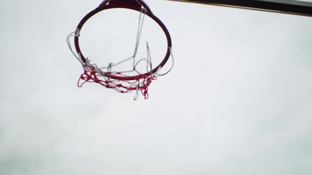 worn basketball hoop against cloudy sky, tattered net swaying empty court evokes memory of street player practicing alone, coach whistle absent, child dreaming of shot, twilight training session,