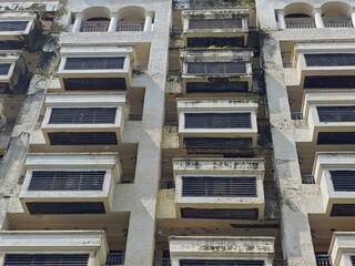 A low-angle shot of a tall, aged residential building featuring repetitive rows of balconies with dark mesh screens and visible weathering on the concrete exterior