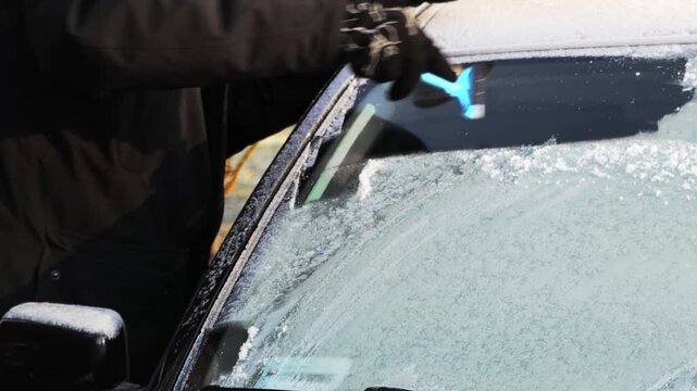 Individual in black coat uses a blue ice scraper to remove frost from a car windshield, demonstrating a continuous action of clearing ice for visibility and safety