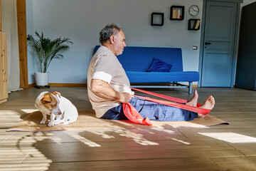 Mature man doing fitness training with resistance band on fitness mat with his Jack Russell Terrier dog at home. Healthy lifestyle for wellness. © Caterina Trimarchi