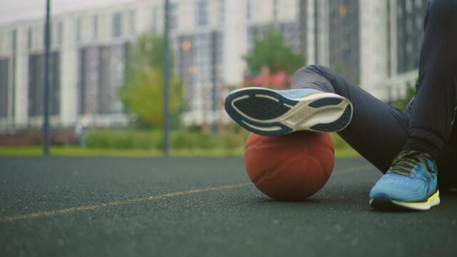 athlete on court with basketball balancing while sitting on asphalt near apartment blocks wearing blue sneakers and dark joggers practicing ball control and leg coordination lowangle cinematic frame