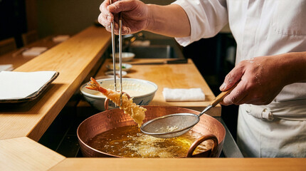 Close-up of Japanese tempura chef's hands frying shrimp in a copper pot at a wooden counter