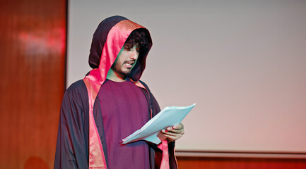 Indian young adult gen z male man standing wear outfit dress hold script paper do play scenes practice indoor hall stage. Teen age boy guy read story line talk speak day time college campus room place