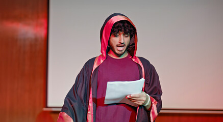 Indian young adult gen z male man standing wear outfit dress hold script paper do play scenes practice indoor hall stage. Teen age boy guy read story line talk speak day time college campus room place