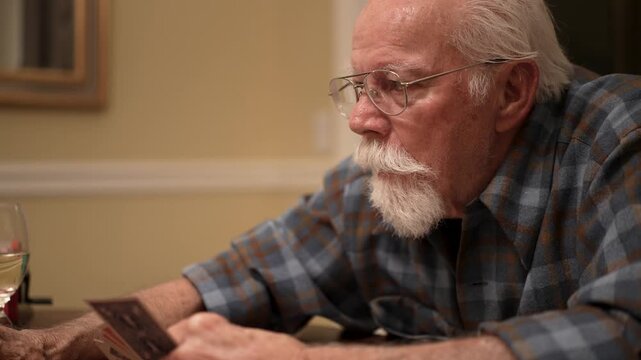 An elderly senior man plays cards alone at his home during the evening, focused on his game with a drink nearby.