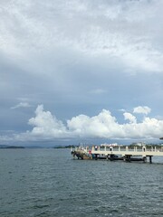 Obraz premium Pier structure extending into calm waters with cityscape in the background and dramatic cumulus clouds overhead. Captures the contrast between nature and urban infrastructure.
