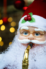 Vertical shot of  toy Santa Claus playing a trumpet surrounded by festive lights and Christmass balls . Close up view