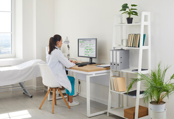 Doctor in clinic office typing on computer. The doctor reviews records on the monitor at desk, using software to manage patient data efficiently. Concept image of digital healthcare workflow.