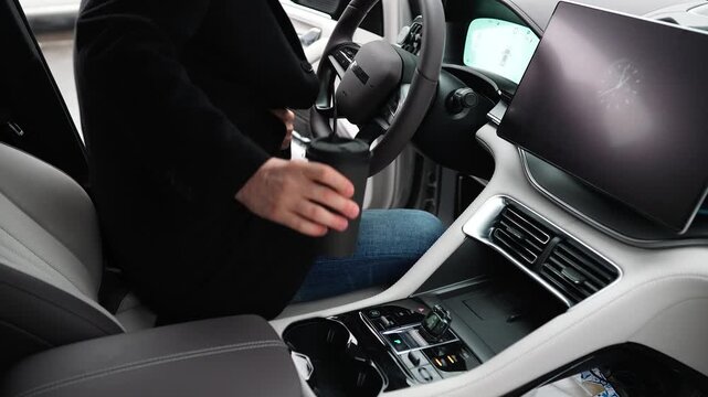Businessman getting into car. Male driver in coat gets into electric car, puts coffee in cup holder and closes the door. The road home after a productive day at work. View through the passenger door.