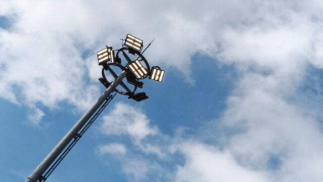 High mast lighting pole with multiple LED floodlights against a bright cloudy sky background.