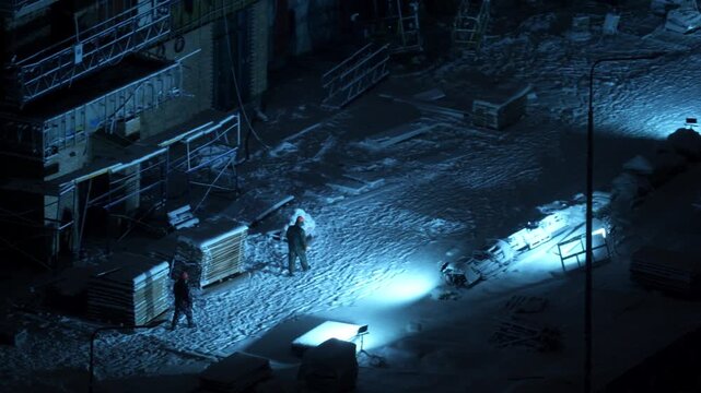 Heavy snowfall during winter night near urban construction site. Snow falling under artificial light, silhouettes of people walking, cold winter weather conditions, outdoor city environment