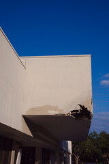 A Damaged cracked exterior wall and damaged overhang beneath a building, with loose debris The scene conveys hazard, neglect, and the vulnerability of urban structures.
